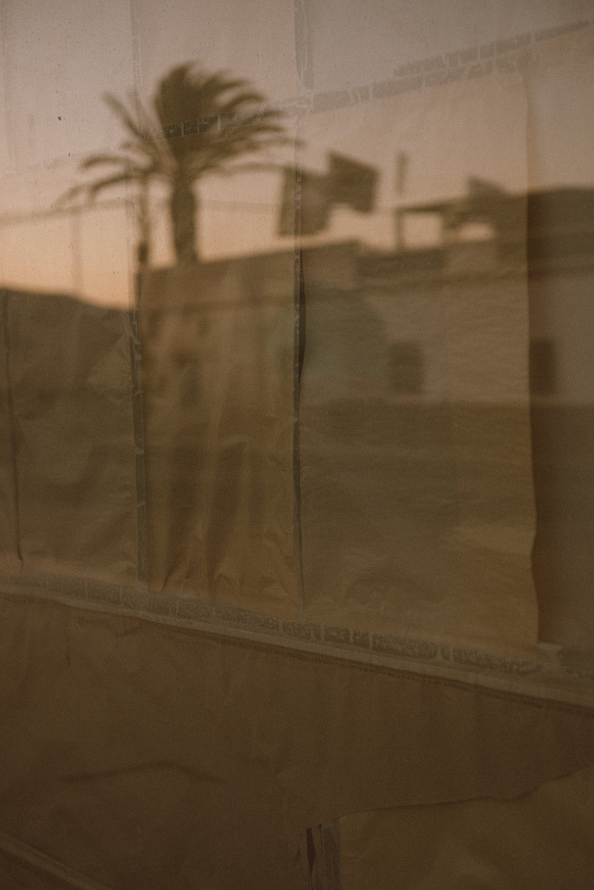 A sepia-toned photograph showing a draped beige fabric or paper sheet with soft folds, overlaid on a warm brown background with a faint reflection of a palm tree and buildings.